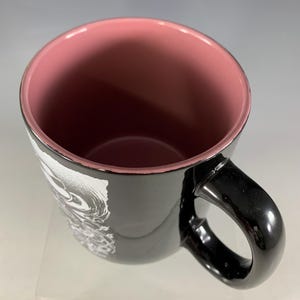 K&ouml;nnte beinhalten: Schwarze Keramik-Tasse mit rosa Innenraum und schwarzem Henkel. Das &Auml;u&szlig;ere zeigt ein wei&szlig;es und graues grafisches Design. Die Tasse ist leer und von oben betrachtet, wobei die Innenfarbe zur Geltung kommt.