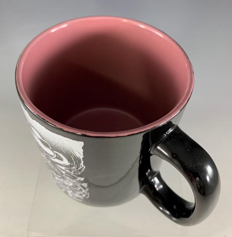 K&ouml;nnte beinhalten: Schwarze Keramik-Tasse mit rosa Innenraum und schwarzem Henkel. Das &Auml;u&szlig;ere zeigt ein wei&szlig;es und graues grafisches Design. Die Tasse ist leer und von oben betrachtet, wobei die Innenfarbe zur Geltung kommt.