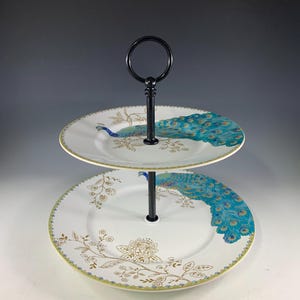 May include: Two-tiered serving stand with white plates featuring peacock and floral designs in gold and teal. The plates have a gold rim and are connected by a black metal handle. Ideal for serving desserts or appetizers.