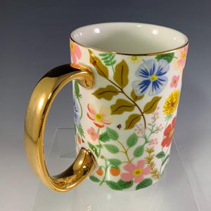 Może przedstawiać: Biały ceramiczny kubek ze złotym uchwytem i obramowaniem, ozdobiony kolorowym kwiatowym wzorem. Wz&oacute;r zawiera r&oacute;żowe, niebieskie, ż&oacute;łte i czerwone kwiaty, a także zielone liście i biedronki. Dekoracyjny kubek.