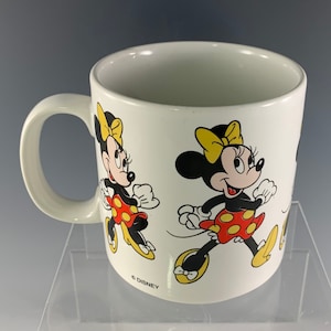 Puede incluir: Taza de cer&aacute;mica blanca con asa que presenta una imagen de dibujos animados de Minnie Mouse con un vestido rojo y blanco de lunares y un lazo amarillo. La taza tiene el texto "&copy; DISNEY" en la parte inferior.