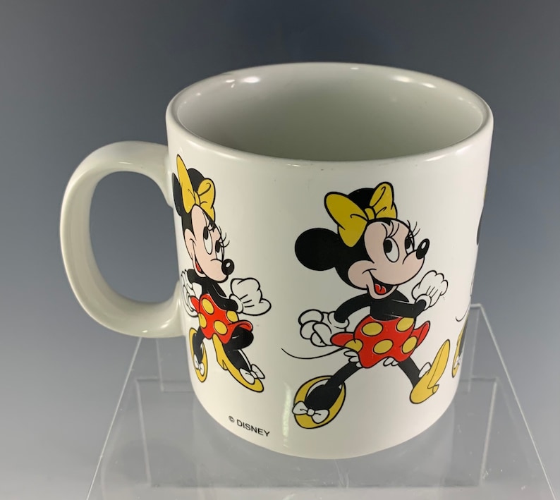 Puede incluir: Taza de cer&aacute;mica blanca con asa que presenta una imagen de dibujos animados de Minnie Mouse con un vestido rojo y blanco de lunares y un lazo amarillo. La taza tiene el texto "&copy; DISNEY" en la parte inferior.