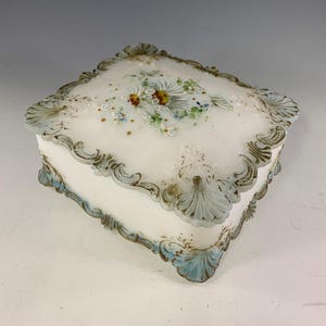 May include: A rectangular white milk glass box with a hinged lid. The lid features a hand-painted floral design with daisies and green foliage. The edges of the lid and base are decorated with raised, scalloped relief patterns in blue and gold.