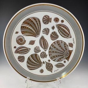May include: A white ceramic plate with a gold rim and a gray center. The plate is decorated with a pattern of stylized seashells in shades of brown and gold. The text "Jorgen Gull" is written in gold on the bottom right corner of the plate.