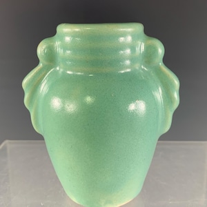 May include: A light green ceramic vase with a rounded body and a wide opening. The vase has two decorative handles on either side and a textured surface. The vase is approximately 6 inches tall.