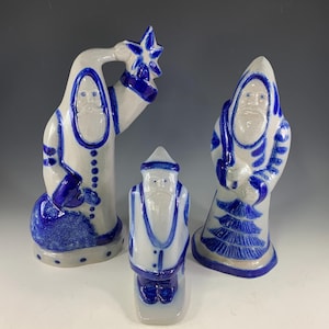 May include: Three ceramic figurines featuring a white base with cobalt blue painted details. One figure holds a star, another carries a sack, and the third holds a small tree. Each figure has a bearded face and a pointed hat, finished with a glossy glaze.