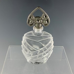 May include: A clear glass bottle with a decorative, heart-shaped, silver-toned stopper featuring floral accents. The bottle has a faceted design.