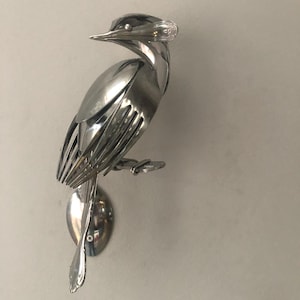 Op de afbeelding: Handgemaakte metalen vogelsculptuur gemaakt van hergebruikt zilverwerk. De vogel zit op een lepelbasis, met vorken die de vleugels en het lichaam vormen. Een uniek kunstwerk voor huisdecoratie, gemaakt van hergebruikt bestek.