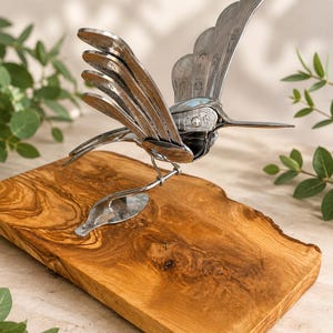 Könnte beinhalten: Eine Kolibri-Skulptur aus silberfarbenem Metallbesteck mit geschichteten Löffelschalen als Flügeln und einem Gabelgriff als Schnabel, montiert auf einem polierten Holzsockel mit natürlicher Maserung und unregelmäßigem, naturbelassenem Rand.