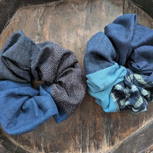 May include: Two handmade scrunchies in shades of blue and black. One scrunchie features a patchwork design with a mix of solid and patterned fabrics. The other scrunchie has a solid blue base with a contrasting blue and patterned fabric.