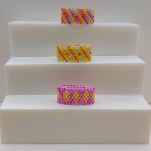 Op de afbeelding: Drie kralenringen in roze, geel en oranje worden getoond op een wit, trapsgewijs platform. De ringen hebben ingewikkelde patronen, waarbij de bovenste ring een gestreept ontwerp heeft en de andere twee chevronpatronen.