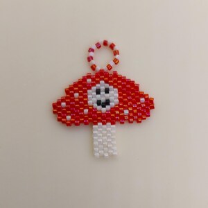 Op de afbeelding: Een rode en witte kralen paddenstoel bedel met een glimlachend gezicht. De bedel heeft een lus aan de bovenkant om aan een sleutelhanger of ander item te bevestigen.