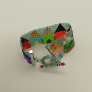 May include: A colourful beaded bracelet with a geometric pattern and a silver clasp. The bracelet is made of small, colourful beads that are arranged in a repeating pattern of triangles. The clasp is a simple, silver toggle clasp.