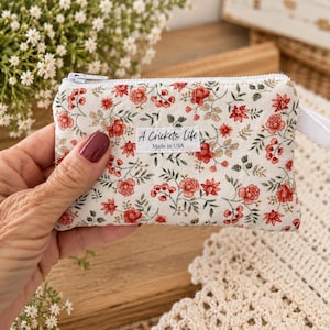 May include: A small rectangular fabric coin purse featuring a cream background with a red and green floral pattern. The pouch has a white zipper closure, a white side tab, and a white fabric label reading A Crickets Life Made in USA.