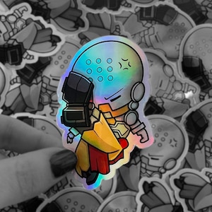 May include: A holographic sticker featuring a cartoon character with a large head, wearing a helmet and a yellow and red outfit. The character has a serious expression and is holding a gun.