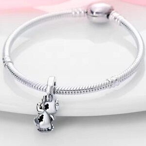 May include: A silver charm bracelet with a small, detailed charm shaped like a cartoon character. The charm has black accents and is suspended from the bracelet. The bracelet is made of a flexible, snake-like chain.