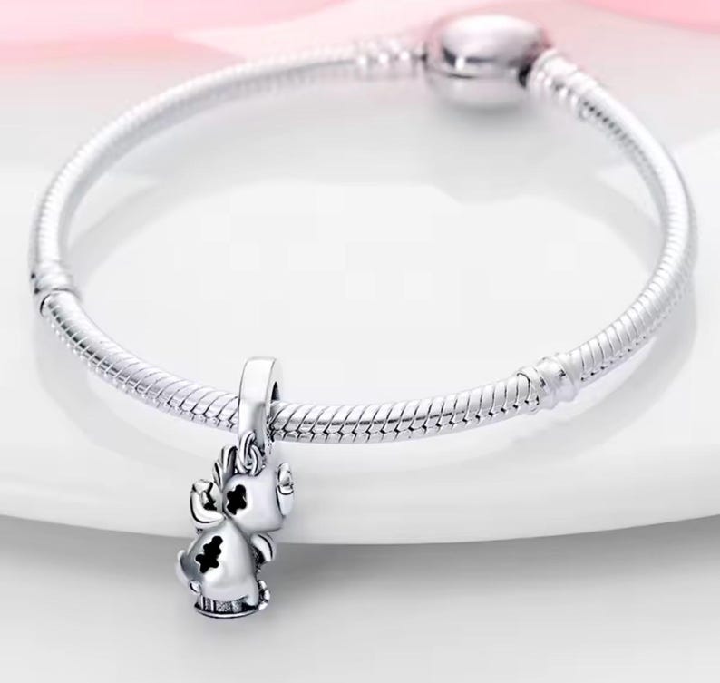 May include: A silver charm bracelet with a small, detailed charm shaped like a cartoon character. The charm has black accents and is suspended from the bracelet. The bracelet is made of a flexible, snake-like chain.