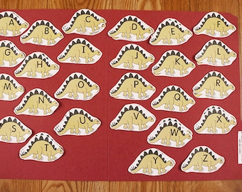 May include: A set of 26 alphabet flashcards featuring dinosaurs. Each card has a yellow dinosaur with black spines and feet, and a black letter printed on it.
