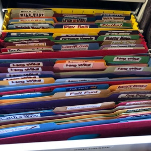 May include: A close-up of a file cabinet drawer filled with colorful file folders labeled with various educational themes, such as "Wild West", "Trick or Treat", "Play Ball", "Octopus Vowels", "Monster Money", "Hog Wild", "Fish Facts", "Fall Fun", "Cookie Antonyms", "Catch a Wave", and "Adding Pencils". The drawer is labeled "2nd Grade".