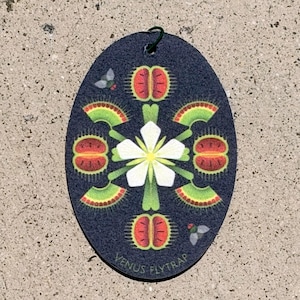 May include: An oval-shaped dark blue air freshener featuring a central white flower surrounded by green and red Venus flytrap illustrations and two small grey flies. The text VENUS FLYTRAP is printed in small light-colored letters at the bottom.