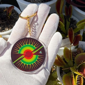 May include: A circular acrylic keychain featuring a vibrant graphic of a Venus flytrap with a red center, yellow and green radiating petals, and a small fly illustration. The keychain includes a gold-toned metal ring and chain attachment.