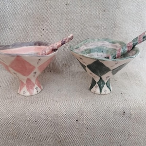 May include: Two ceramic bowls with matching spoons. One bowl is pink and white with a diamond pattern, the other is green and white. Both have a flared shape and are decorated with small floral accents.