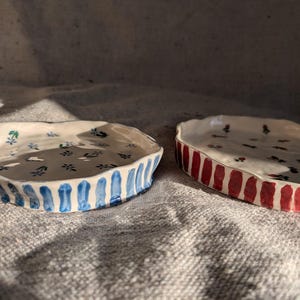 May include: Two handmade ceramic dishes. One is white with blue floral accents and a blue vertical stripe pattern around the edge. The other is white with red vertical stripes and small black floral accents.