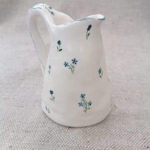 May include: A small, white ceramic jug with a curved handle and a slightly ruffled rim. The jug is decorated with small, blue floral sprigs. The rim and handle are outlined with a thin blue line. The jug is approximately 13 cm tall.