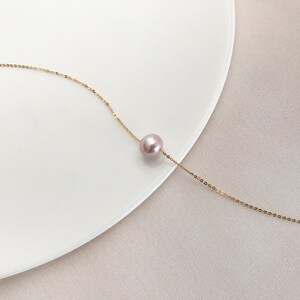 May include: A fine gold chain necklace featuring a single, round, lavender-pink pearl pendant. The necklace is presented on a white surface, against a soft, neutral backdrop.
