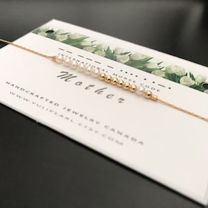 May include: A gold chain necklace with white pearls and gold beads spelling out "Mother" in International Morse Code. The necklace is displayed on a white card with green floral designs and the text "HANDCRAFTED JEWELRY CANADA WWW.YULIPEARL.ETSY.COM".
