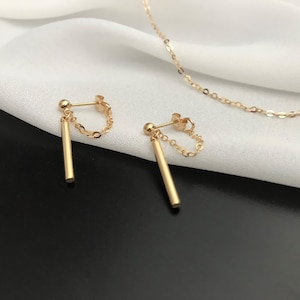 May include: Gold-coloured earrings featuring a post, chain, and vertical bar design. A fine gold necklace is also present. The jewellery is set against a white and black backdrop.