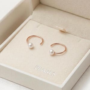 May include: Two rose gold open hoop earrings, each with a single white pearl, displayed in an open white box. The box is labelled "PEARLS" inside. The earrings are a delicate jewellery item.
