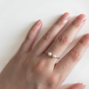 May include: A delicate gold ring with two white pearl accents. The ring is worn on a finger of a hand.