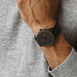 May include: A wristwatch with a black face and black band. The watch has a minimalist design and is shown on a person's wrist. The person is wearing a gray sweater and a brown suede jacket is draped over their shoulder.
