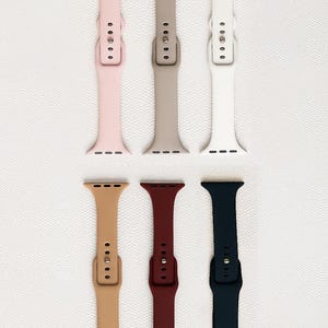 May include: A collection of six watch bands in various colors, including pink, tan, white, light brown, burgundy, and navy blue. Each band features a buckle and multiple adjustment holes for a secure fit. The bands are displayed on a textured white surface.