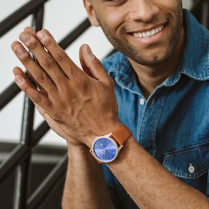 K&ouml;nnte beinhalten: Ein Mann tr&auml;gt ein blaues Jeanshemd und eine schwarze Jeans mit Destroyed-Details. Er tr&auml;gt eine Uhr mit einem braunen Lederarmband und einem blauen Zifferblatt.