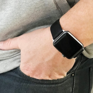 Pode incluir: Um smartwatch com caixa retangular prateada e pulseira de silicone preta lisa. O mostrador &eacute; escuro e inativo, apresentando um design quadrado com cantos arredondados e um bot&atilde;o de coroa lateral, usado com um moletom cinza e cal&ccedil;a jeans escura.