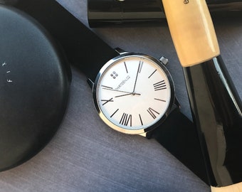 May include: A silver watch with a white face and black leather band, two black compact cases, and a wooden makeup brush with black bristles.