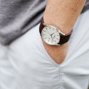 May include: A silver watch with a white face and brown leather band. The watch is on a person's wrist.