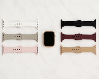 Könnte beinhalten: Eine Apple Watch mit goldenem Gehäuse und schwarzem Bildschirm ist von sechs Armbändern in verschiedenen Farben umgeben: Weiß, Grau, Rosa, Schwarz, Burgunderrot und Beige. Die Armbänder sind aus Silikon und haben einen Schnallenverschluss.