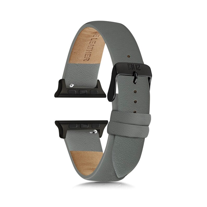 May include: A grey leather watch band featuring a black metal buckle and black attachment hardware. The interior of the band is lined with light tan leather and includes the embossed text "GENUINE LEATHER" on the inner surface of the strap.