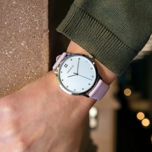 Puede incluir: Un reloj de pulsera plateado con esfera blanca y correa de cuero rosa claro. El reloj tiene un dise&ntilde;o minimalista con peque&ntilde;os marcadores y la marca "SWISS MADE". El reloj se lleva en la mu&ntilde;eca.