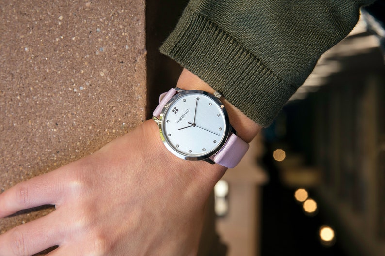 Puede incluir: Un reloj de pulsera plateado con esfera blanca y correa de cuero rosa claro. El reloj tiene un dise&ntilde;o minimalista con peque&ntilde;os marcadores y la marca "SWISS MADE". El reloj se lleva en la mu&ntilde;eca.