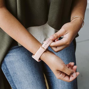 Puede incluir: Una persona ajusta una correa de reloj de cuero rosa claro en su mu&ntilde;eca. La correa tiene una hebilla de oro rosa.