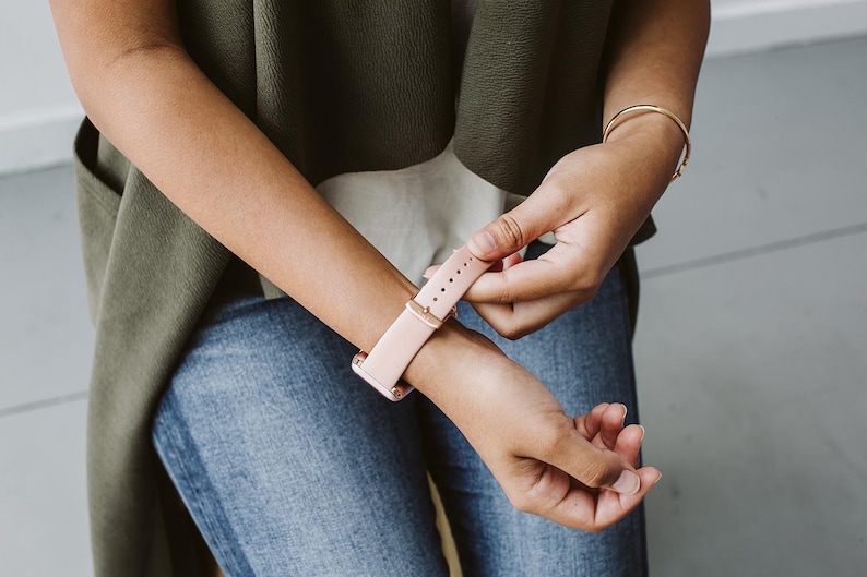 Puede incluir: Una persona ajusta una correa de reloj de cuero rosa claro en su mu&ntilde;eca. La correa tiene una hebilla de oro rosa.