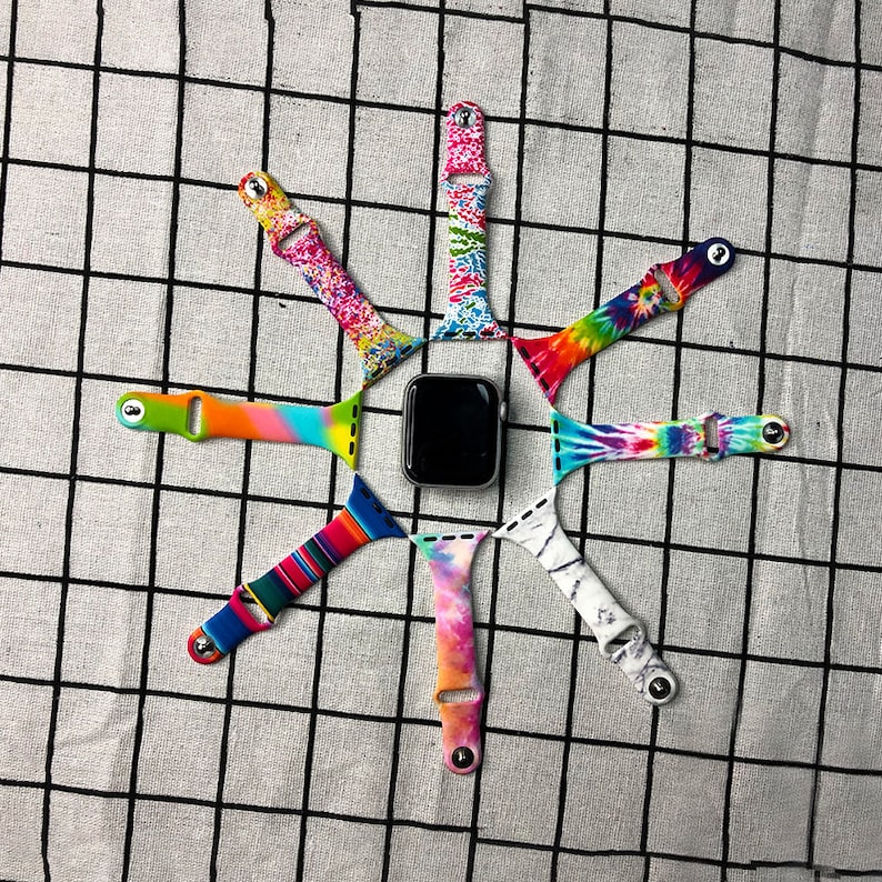 Puede incluir: Ocho correas de reloj de silicona de colores dispuestas en forma de estrella alrededor de un Apple Watch negro. Las correas presentan patrones tie-dye, de m&aacute;rmol y geom&eacute;tricos.
