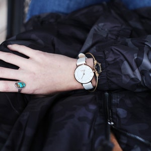 May include: A white wristwatch with a rose gold band and a white face. The watch is worn on a person's wrist with a turquoise ring on the same hand.