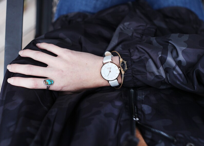 May include: A white wristwatch with a rose gold band and a white face. The watch is worn on a person's wrist with a turquoise ring on the same hand.