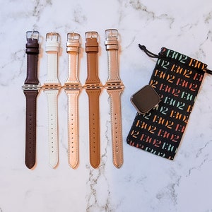 May include: Five different colored Apple Watch bands. The bands are brown, white, rose gold, tan, and gold. The bands are all made of leather and have a silver buckle. The bands are arranged in a row on a white marble surface. A black pouch with white text is in the background.