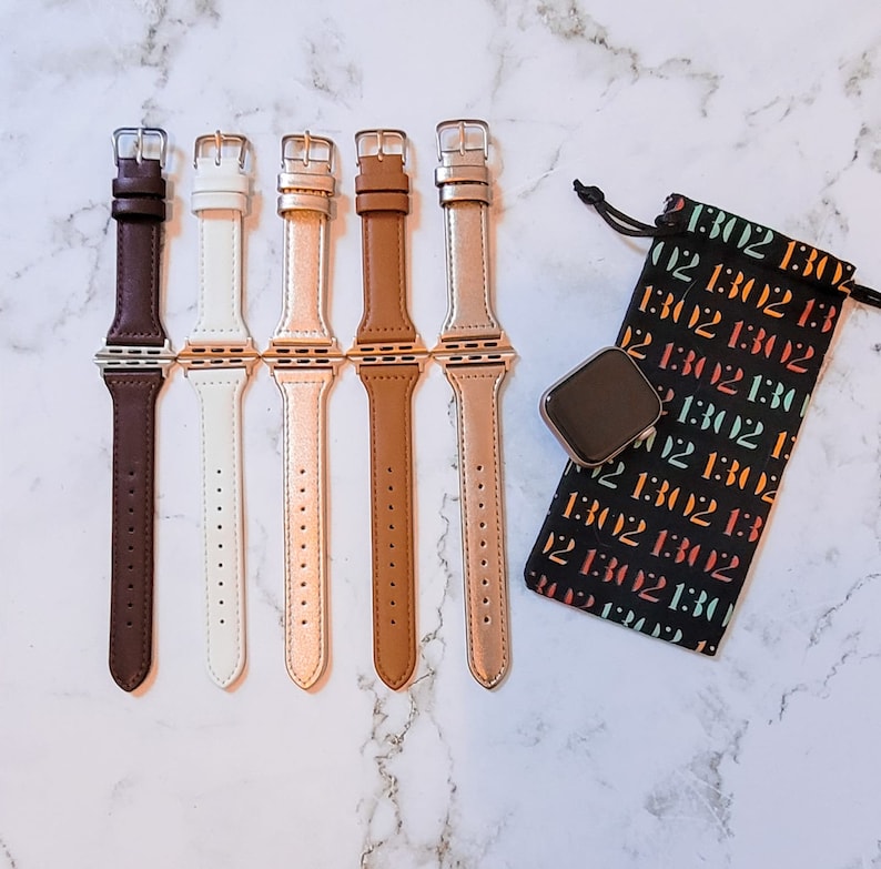 May include: Five different colored Apple Watch bands. The bands are brown, white, rose gold, tan, and gold. The bands are all made of leather and have a silver buckle. The bands are arranged in a row on a white marble surface. A black pouch with white text is in the background.